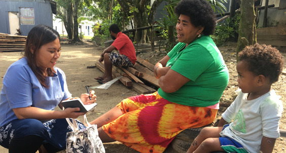 a woman in a blue shirt sits next to two people, wearing colorful clothing. the woman has a notebook and she is listening to the two people speak and taking notes