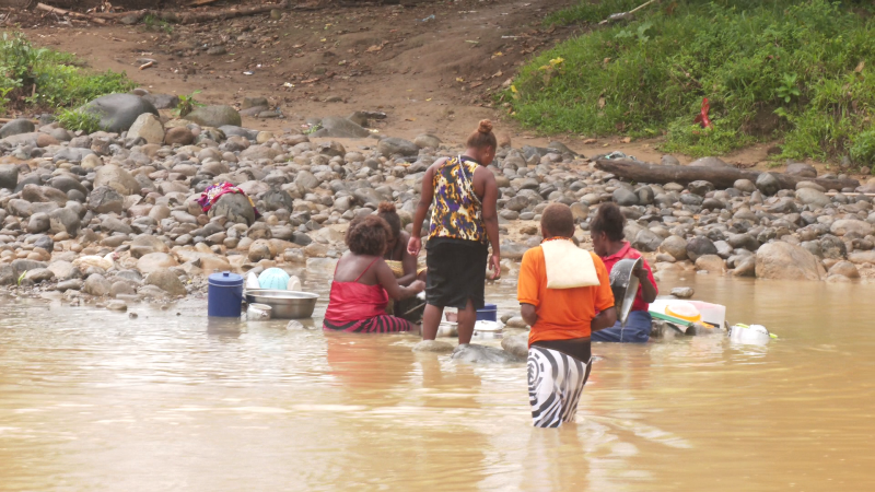 women standing in a yellow, muddy river wearing shawls and doing dishes in the shallow water next to some rocks