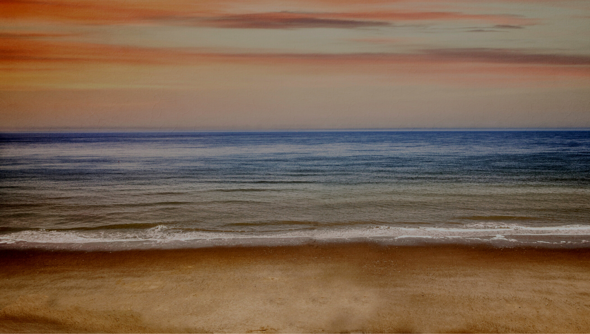 coastal beach view, looking at the waves lapping the shore and the ocean is blue with an orange tint from the sunset in the background