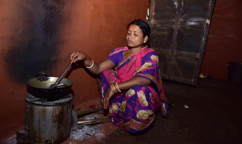 A woman kneels on the floor of a dark room stirring a pot that is being cooked on a solar stove