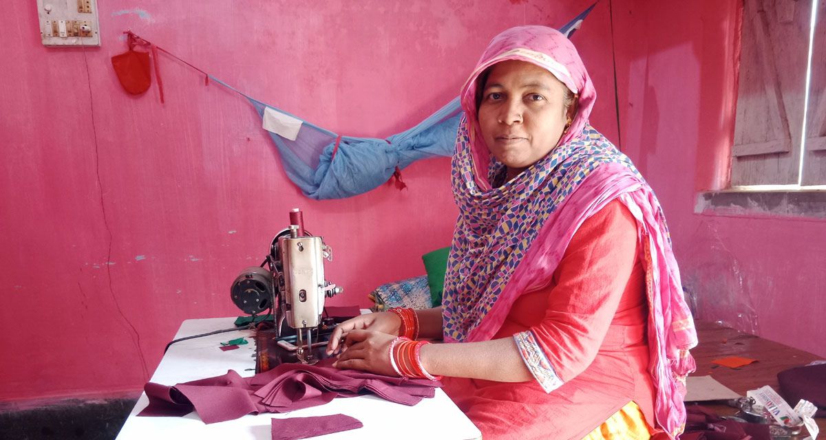 A woman sitting in a room with pink walls, she is wearing a pink and blue spotted shawl over her head and hair, and an orange top that comes to her elbows. she is holding a piece of purple fabric up to a tabletop sewing machine and looking at the camera, seeming pleased