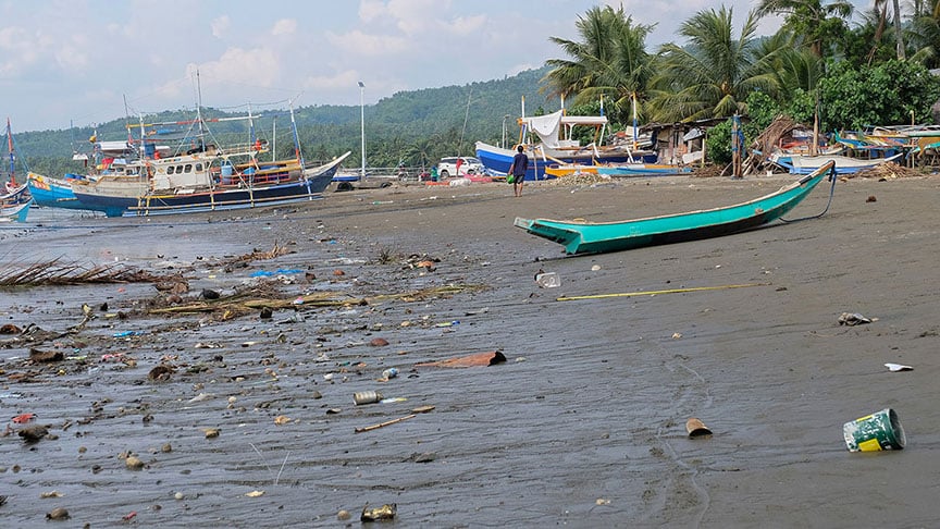 A beach with boats in the background and trash in the foreground. 