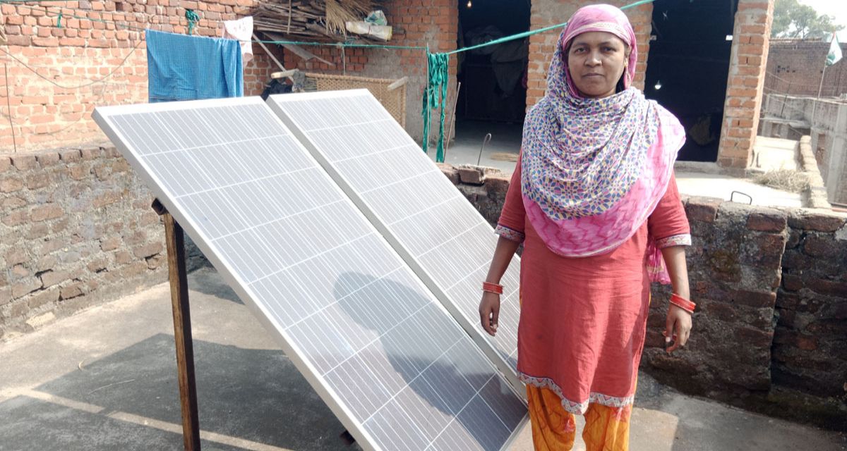 the same woman as the above picture standing in front of a solar panel angled at a 45 degree angle toward the sky, with a red brick house behind her