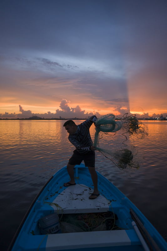 A fisher on a boat throws a net into the ocean at sunset.