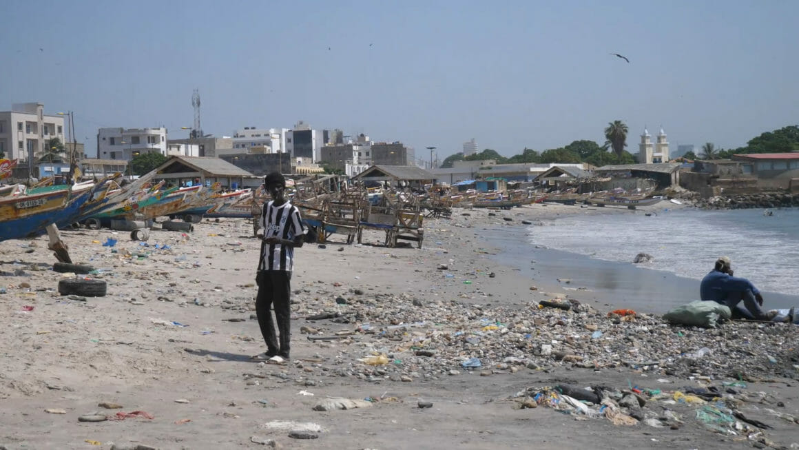 the beach in Senegal. there is trash on the ground, and in the distance you can see houses and fishing boats
