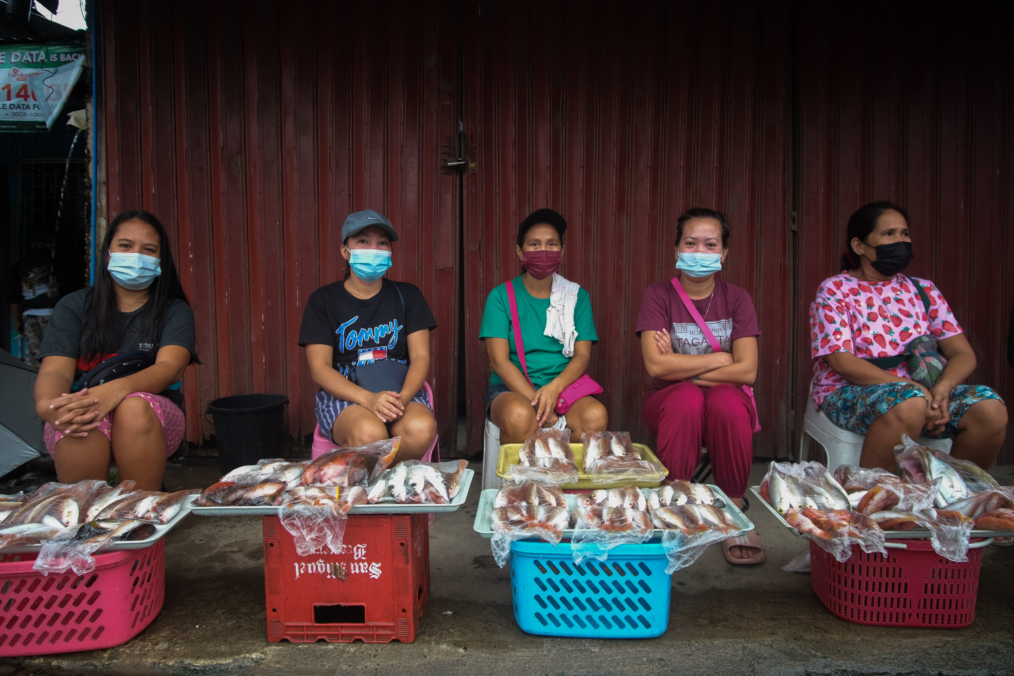 five masked women sit on small stools at a market with buckets and trays of fish lined up in front of them, waiting to sell