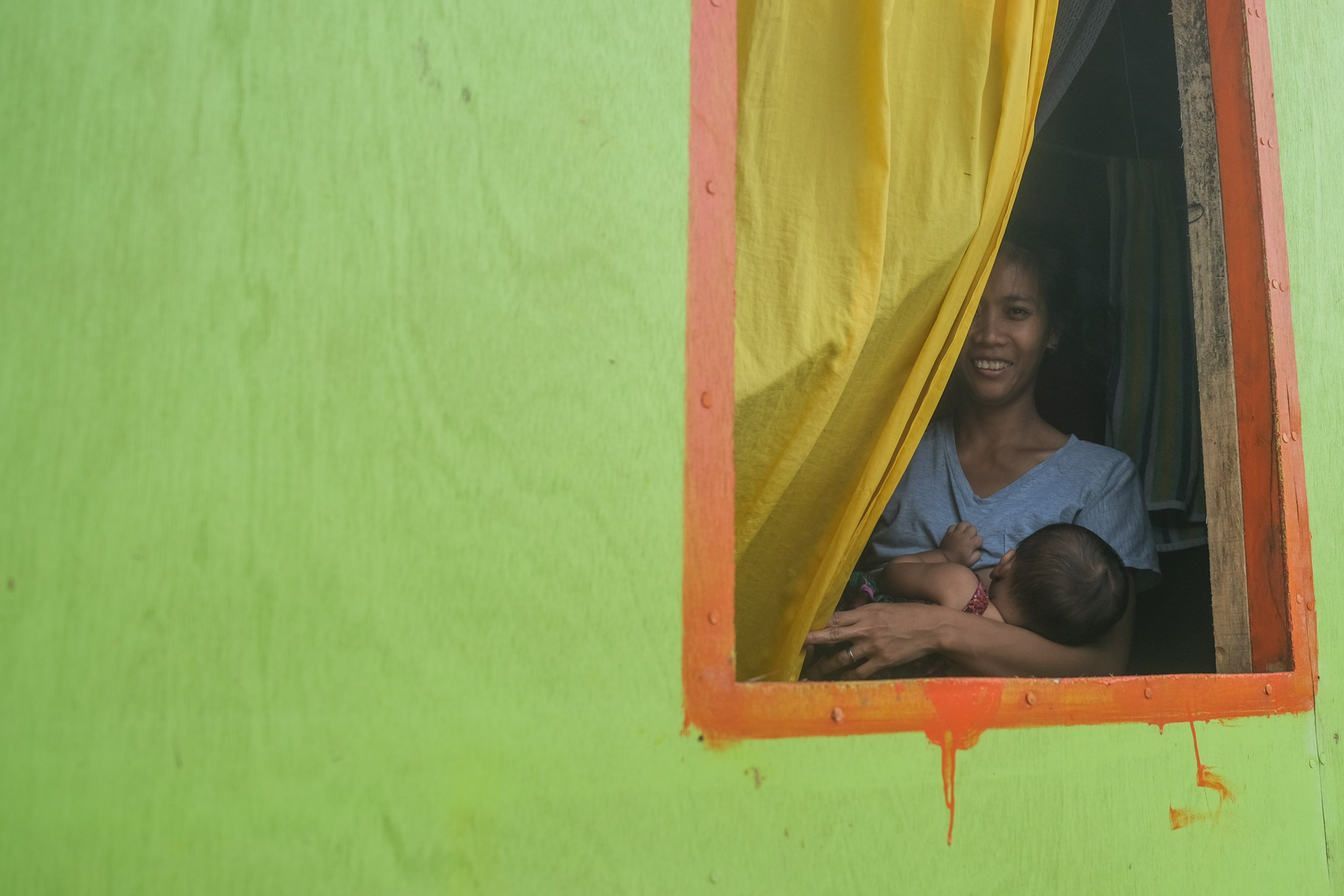 a young mother inside a green house, peering out a window from behind bright yellow curtains. she's holding a baby and breastfeeding as she smiles at the camera