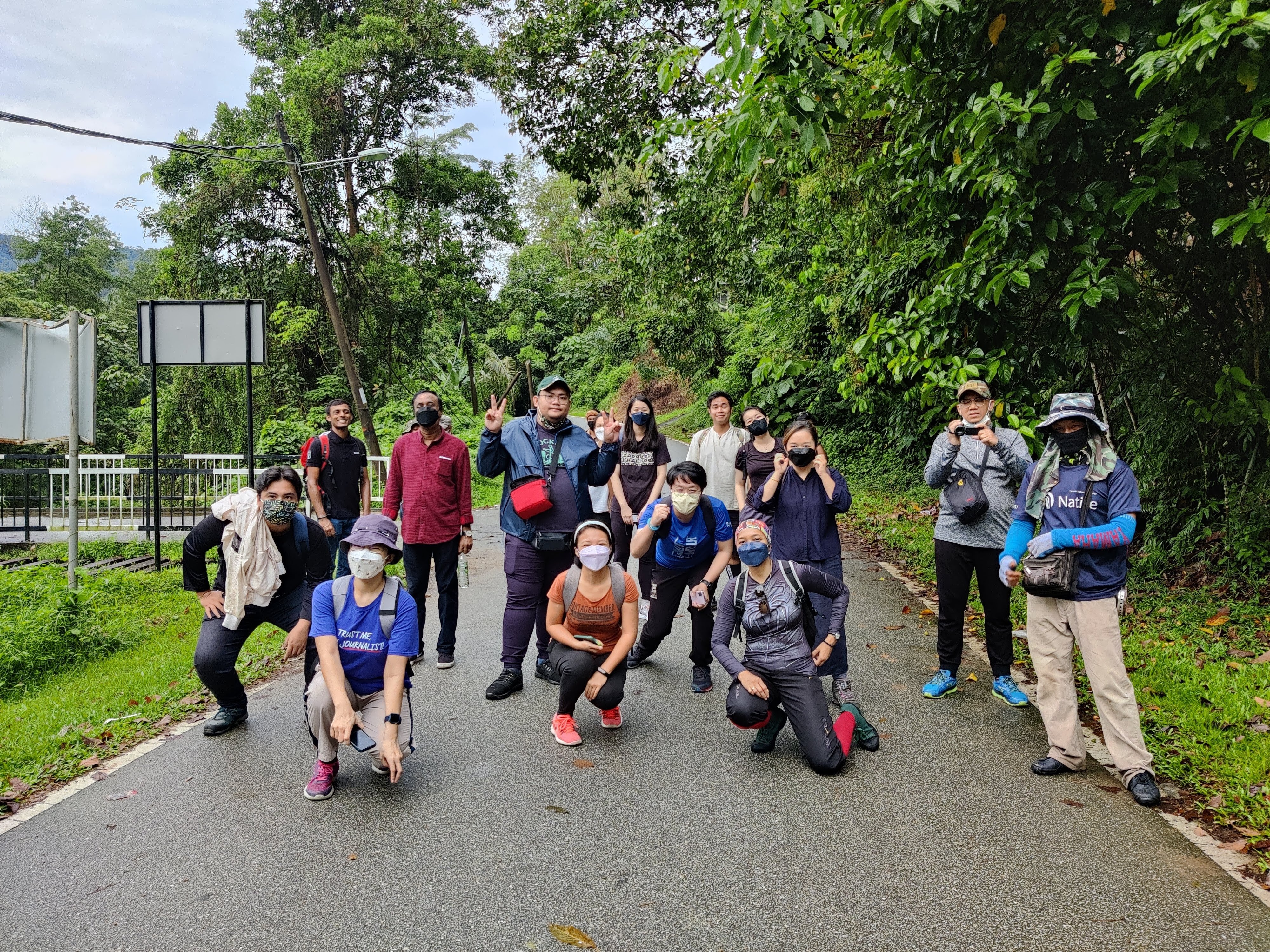 a group of people in hiking attire and gear pose for the camera in a group shot. some of them are masked. they are standing on a path with trees behind them