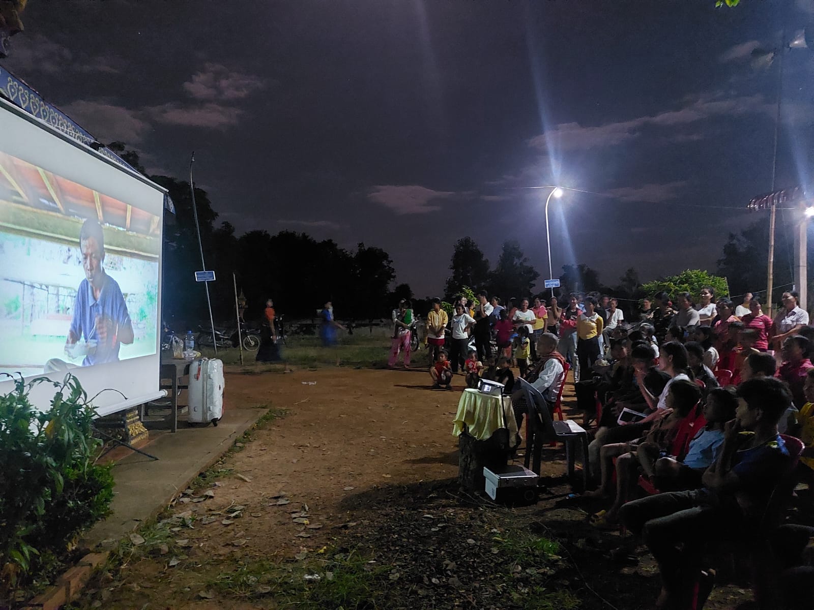 A crowd watches a large movie screen outdoors with trees in the background