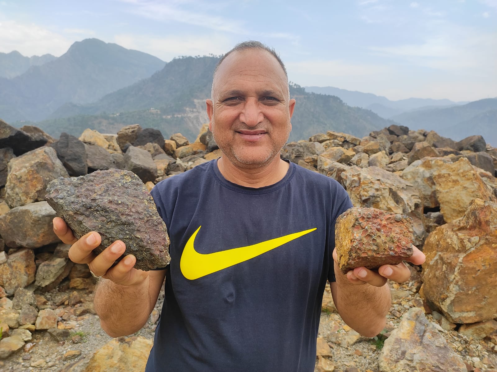 A smiling man, wearing a black shirt with a yellow tick on it, holds up two rocks, one in each hand, as mountains loom in the background.