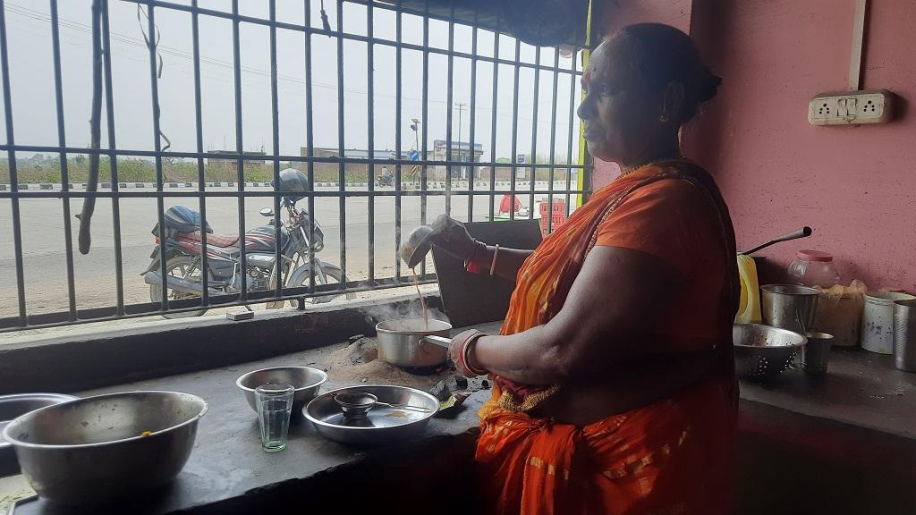 A woman wearing an orange sari smiles slightly as she pours tea looking out of her grilled, kitchen window