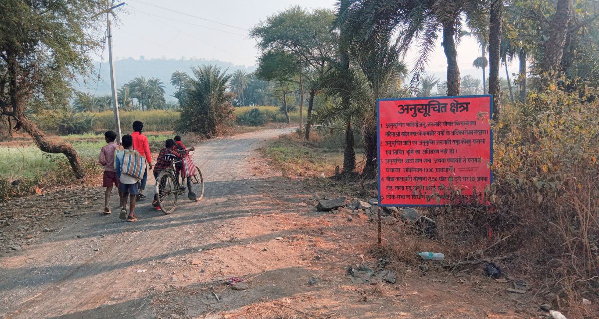 One man and four boys walk down a rural street, as one of them pushes a cycle, past a sign that reads 'Scheduled Area'