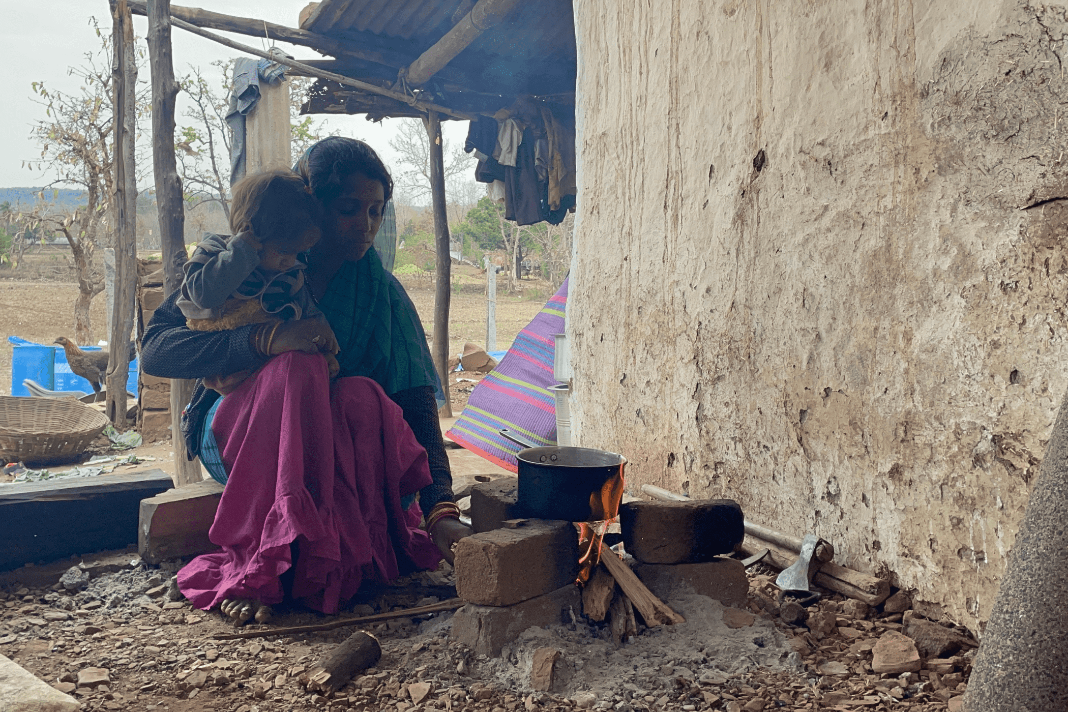 A woman, wearing a pink sari, holds a child in her arms as she adds firewood to the fire that is heating a blackened pot