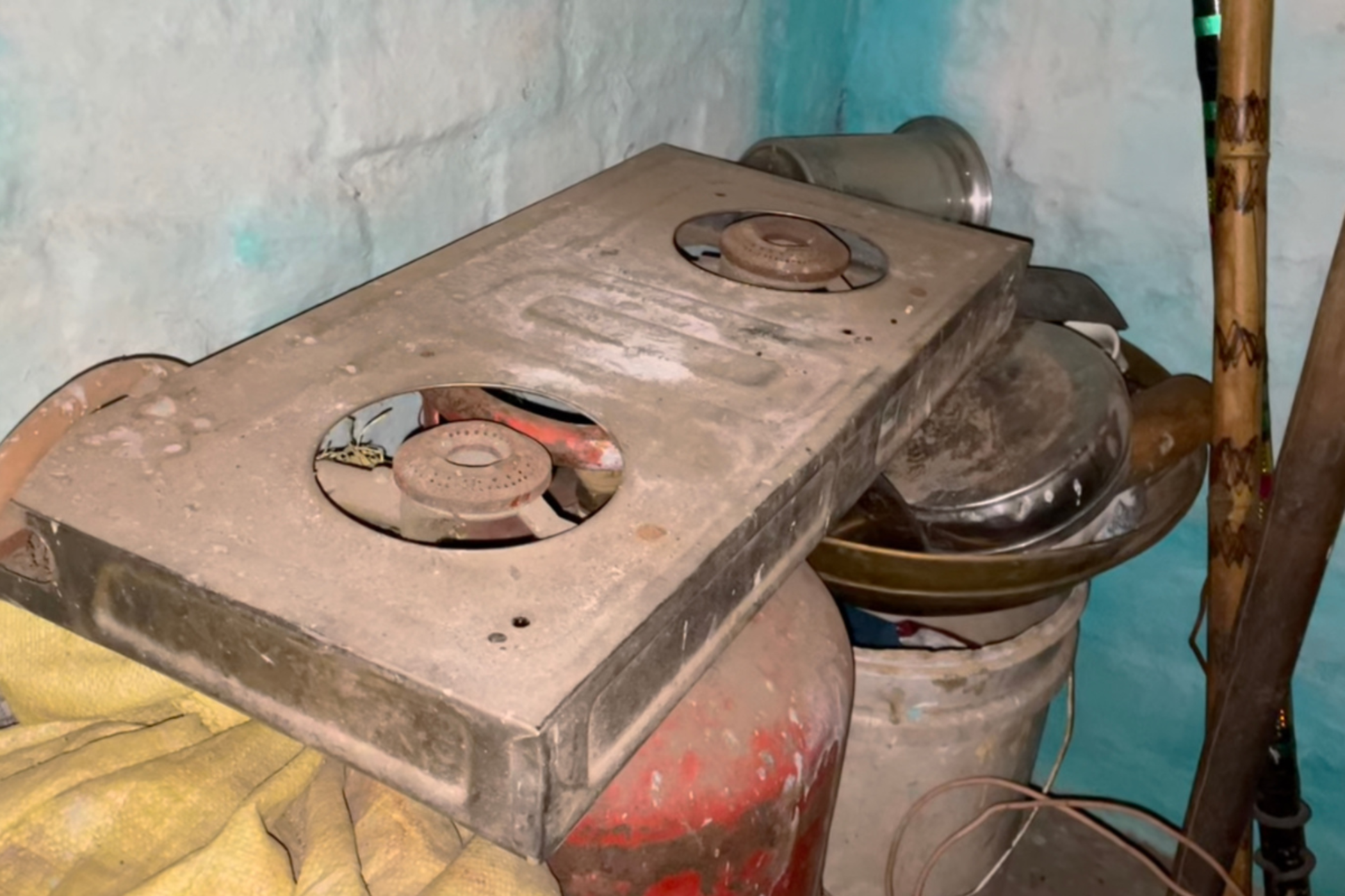 A dusty gas-stove lies on top of a red gas cylinder, a dusty bucket and some overturned vessels.