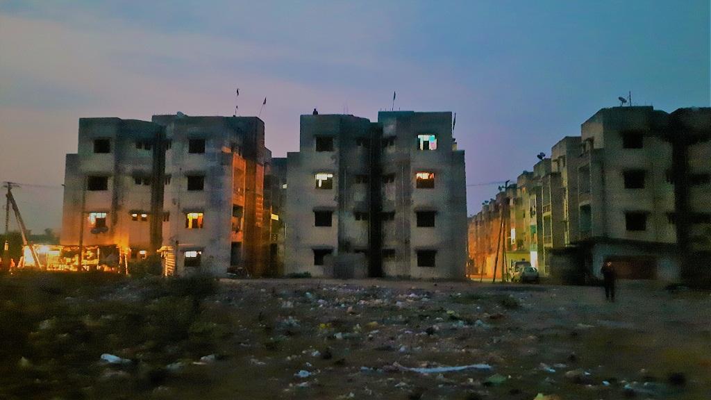 Three storey-buildings with a few lit-up houses overlooks a garbage-strewn field