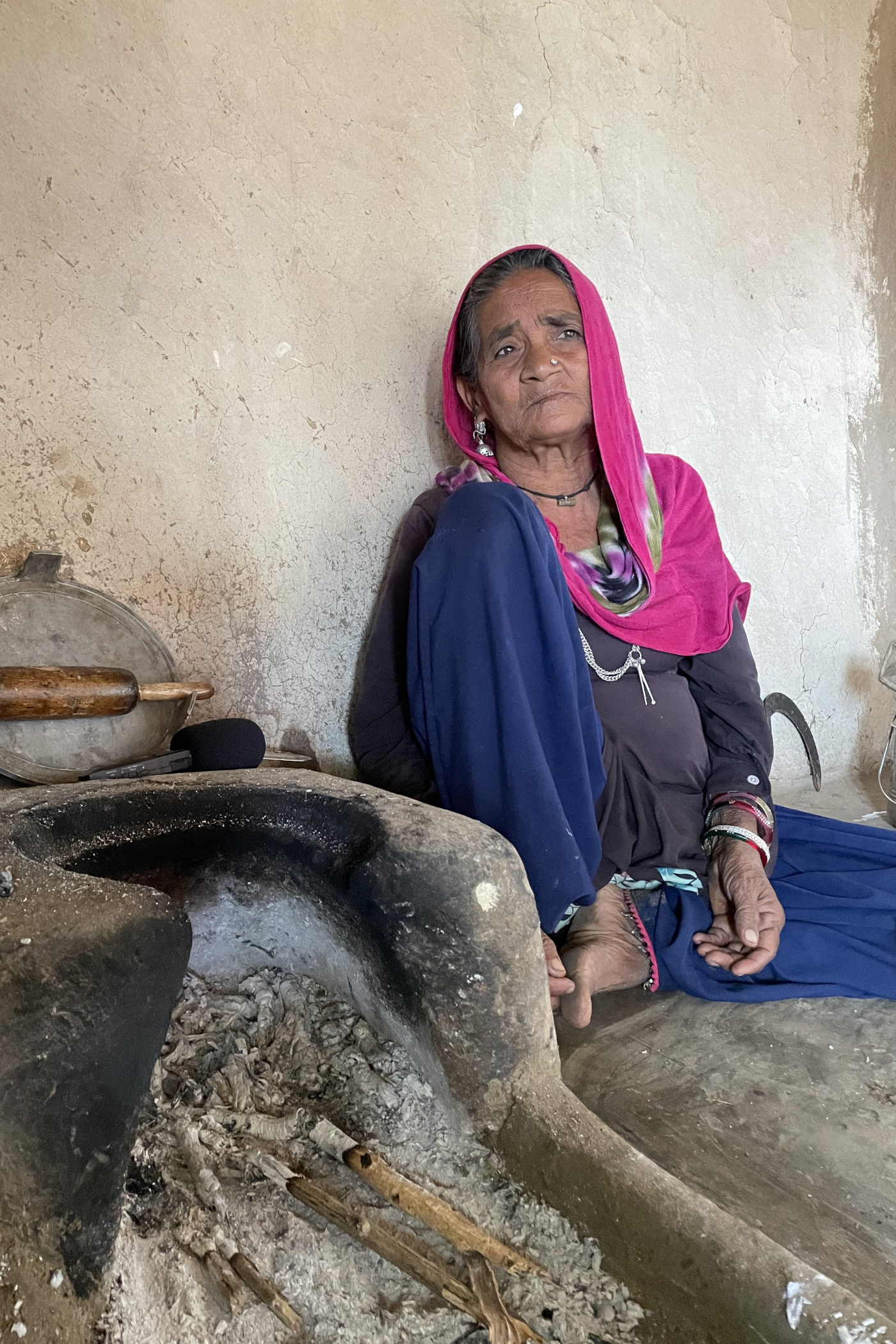 A woman, dressed in a blue salwar-kameez with her head covered by a pink cloth, sits beside an earthen stove which holds ash and half-burned sticks 