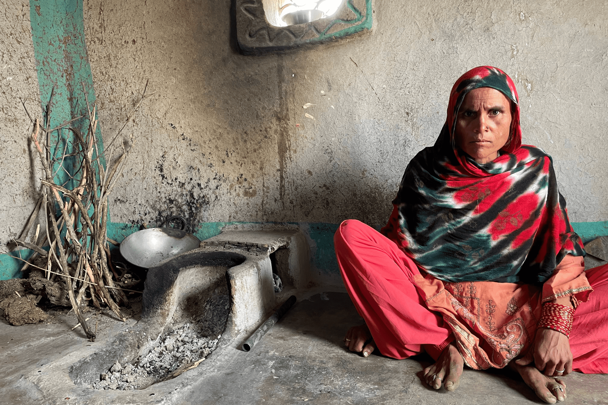 A woman in a red salwar kameez, sits beside an earthen stove and firewood.