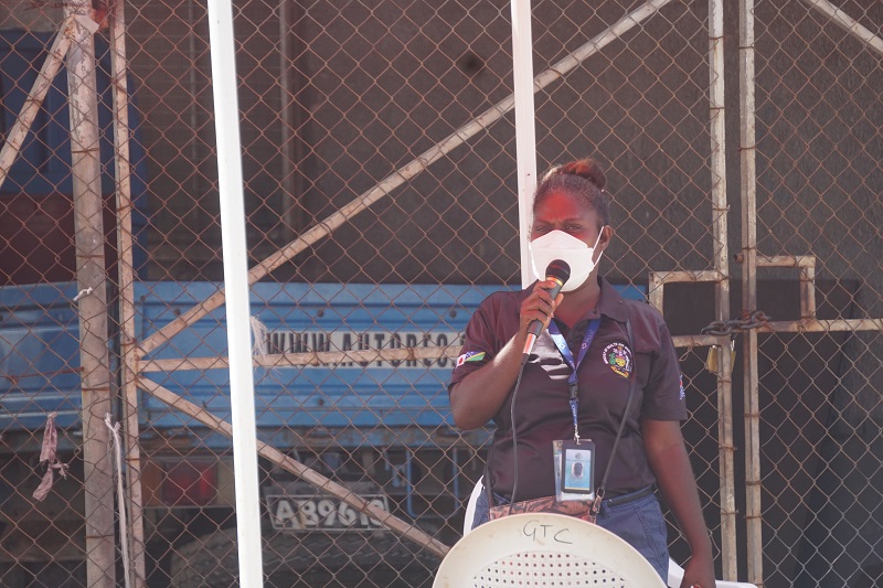 a woman standing in front of a fence looking at the camera, wearing a mask and holding a microphone. she is speaking into it