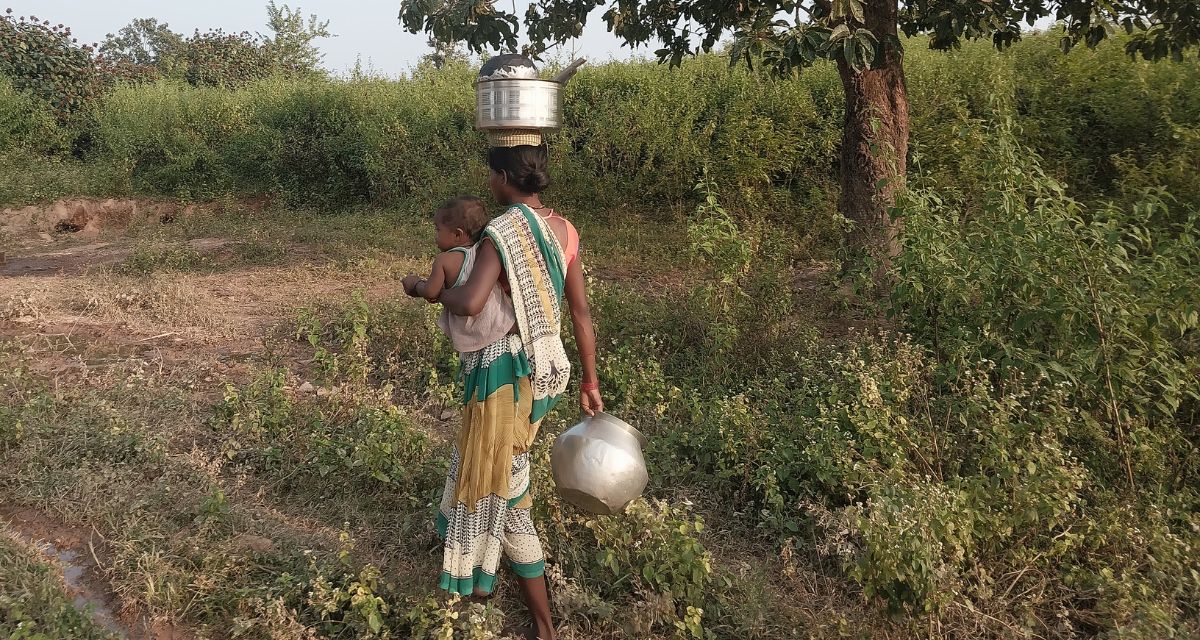A woman holds a baby in one hand and a water jug in another while balancing yet another jug on her head.