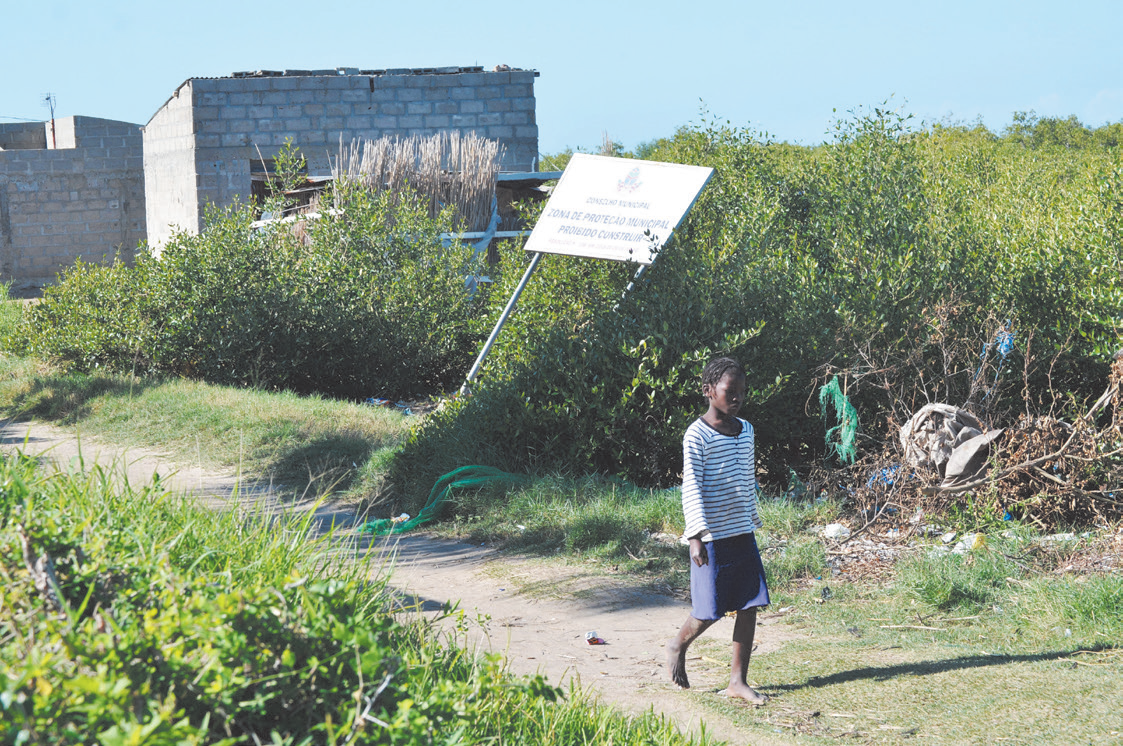 a person walks down the road, behind them there is a building and some trees, and a sign that says "municipal protection zone, prohibited to build" but the sign has fallen over halfway