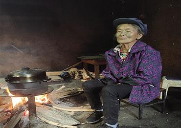 a woman sits next to a fire with a pot cooking wearing a purple/blue jacket and a blue hat, singing