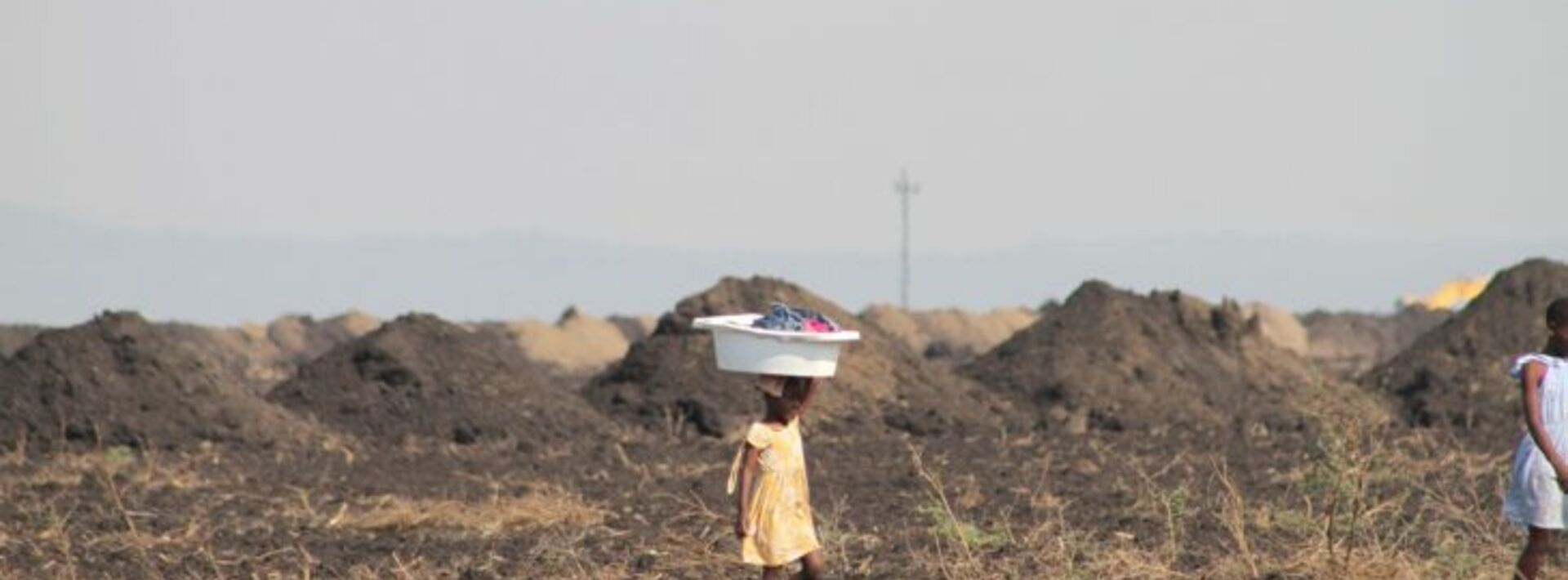 a child carrying a bucket on their head walking through a landscape that is bare and tan without much vegetation