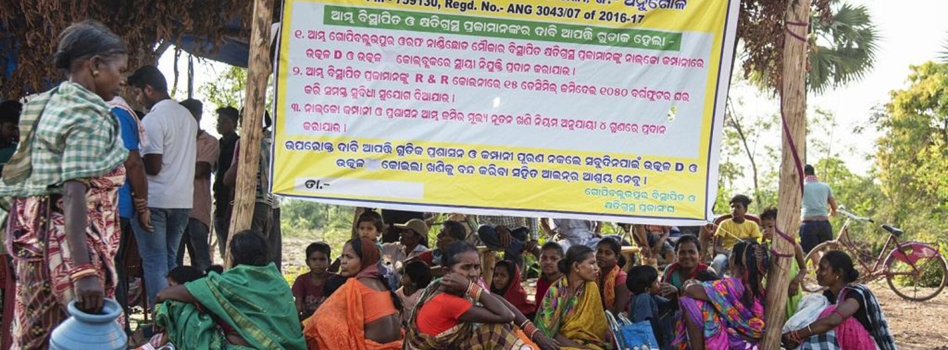 A group of women and children are seated in front of a poster on which a list is presented in Odiya language, while men stand at the back.