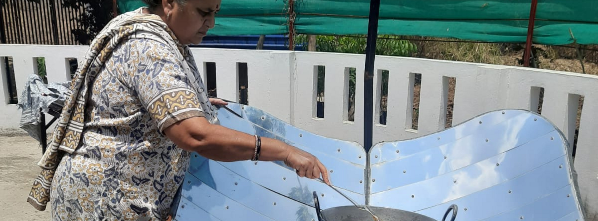 An elderly woman stands on a sunny terrace as she stirs the food in a vessel, resting on a parabolic metal dish.