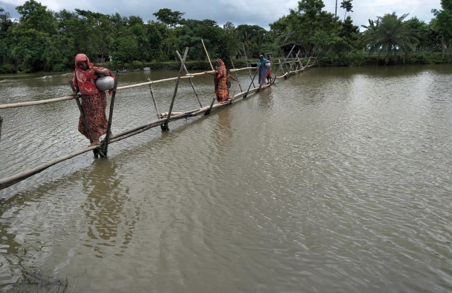Women walking single file carrying pots of water across a flooded area in Bangladesh.