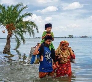 Two adults walking in a flooded area of India's Sundarbans with a child on one person's shoulder, carrying bags