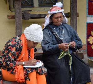 EJN staff member Stella Paul wearing an orange outfit and a hat writing on a notepad while she speaks with an Indigenous women dressed in dark blue who is knitting