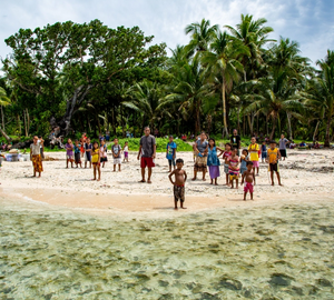 a group of people standing on the shoreline of an island with trees behind the, they are wearing all different clothes and looking at the camera, the person taking the picture is standing in the water facing the island
