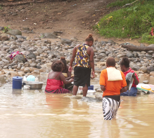 women standing in a yellow, muddy river wearing shawls and doing dishes in the shallow water next to some rocks