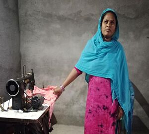 a woman stands in front of grey walls, with a blue shawl across the top of her body and her head. her dress is pink underneath. she stands next to a table with a tabletop sewing machine on it and looks proudly at the camera