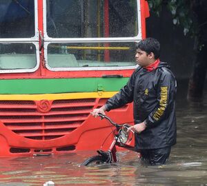 a man holding the handlebars of a bicycle while walking on a road, he is up to his knees in water and is walking in front of a submerged bus