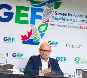 head of gef sitting at a table, black tablecloth, wearing a suit. behind him there is a banner with the gef logo