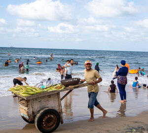 a man with a two wheeler yellow cart walking along a beach with the ocean in the background and many people swimming, he is selling corn
