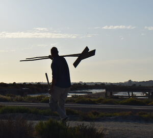 a man carrying tools while walking across a salt marsh with the setting sun in the background on the horizon