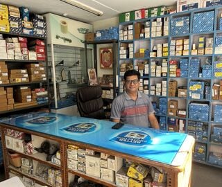 A bespectacled man sits behind a table, on a black chair, and the walls around him are lined with racks full of boxes of different sizes.