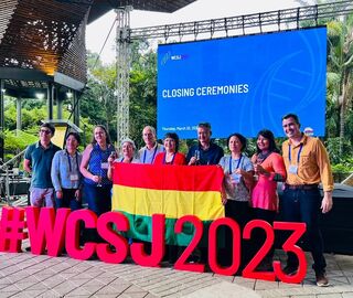 A group of 10 people hold a country flag at an outdoor pavilion; in front of them rest block letters that read "#WCSJ 2023" and behind them is a projector screen that reads "closing ceremonies"