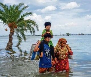 Two adults walking in a flooded area of India's Sundarbans with a child on one person's shoulder, carrying bags