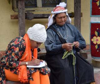 EJN staff member Stella Paul wearing an orange outfit and a hat writing on a notepad while she speaks with an Indigenous women dressed in dark blue who is knitting