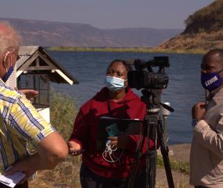 woman interviewing a man with a video camera at a water body