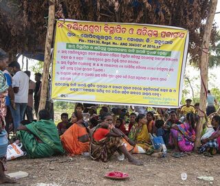 A group of women and children are seated in front of a poster on which a list is presented in Odiya language, while men stand at the back.