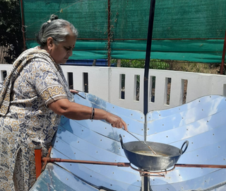 An elderly woman stands on a sunny terrace as she stirs the food in a vessel, resting on a parabolic metal dish.